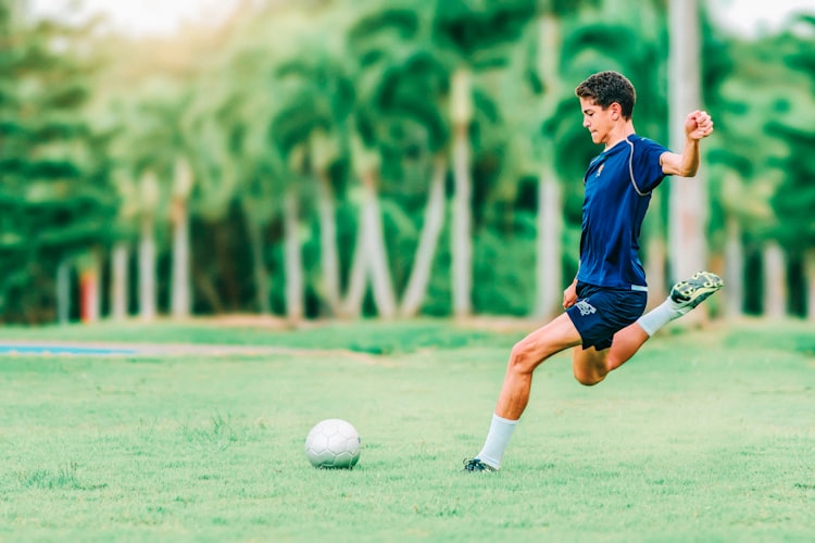 Pemain sepak bola berkaus biru mengayunkan kaki untuk menendang bola di lapangan rumput